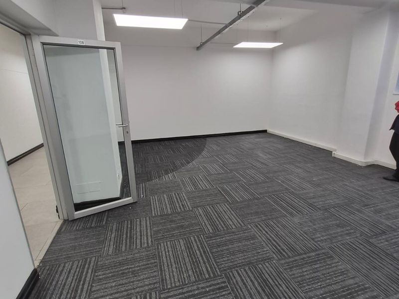 An empty, well-lit office space featuring modern carpet tiles, exposed ceiling conduit, and a glass-paneled aluminum door.