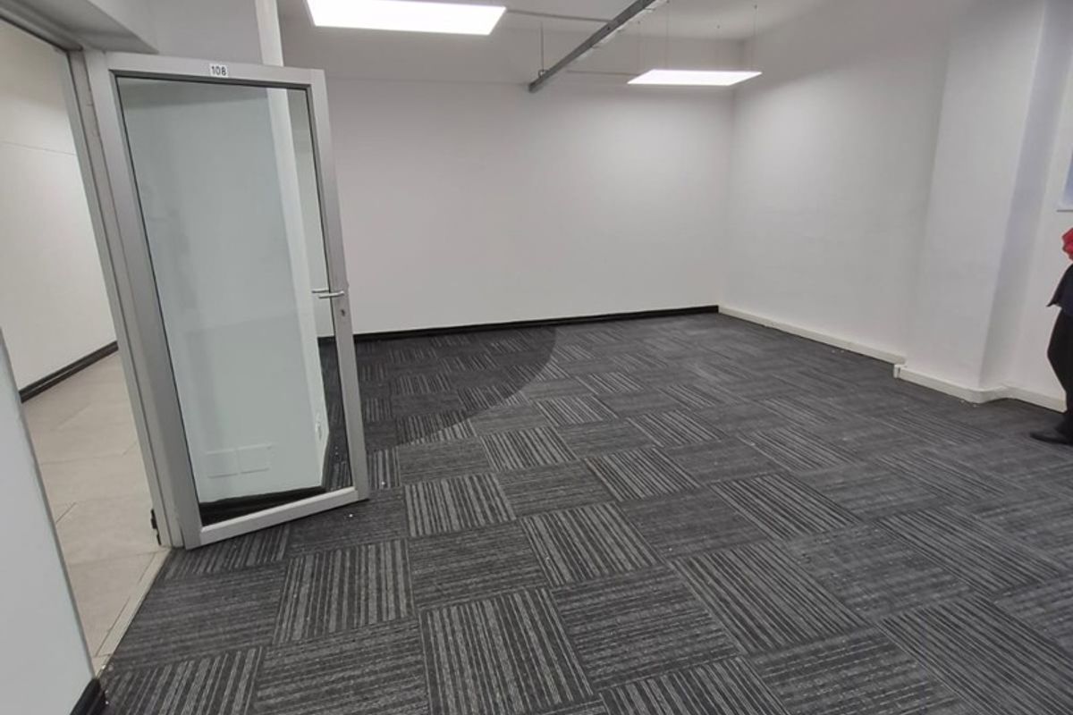 An empty, well-lit office space featuring modern carpet tiles, exposed ceiling conduit, and a glass-paneled aluminum door.