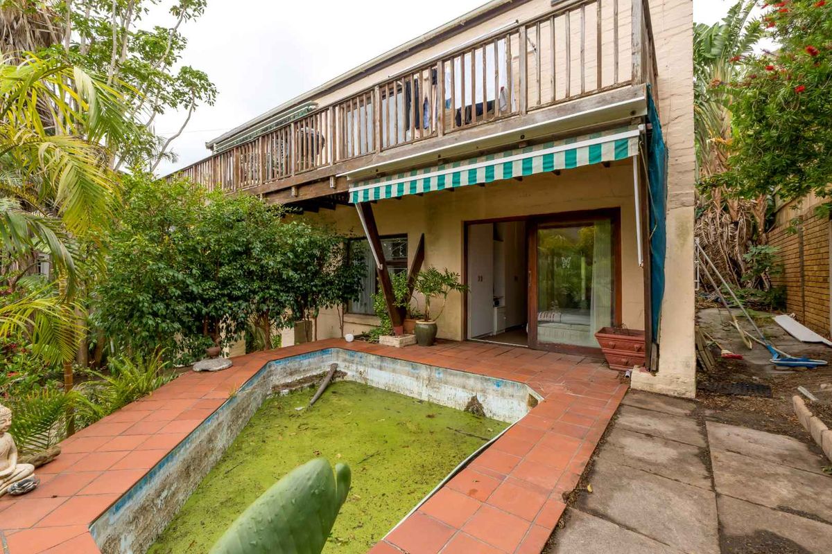 Rear exterior view featuring a neglected swimming pool, paved patio area, and an upper-level wooden balcony with a retractable awning.