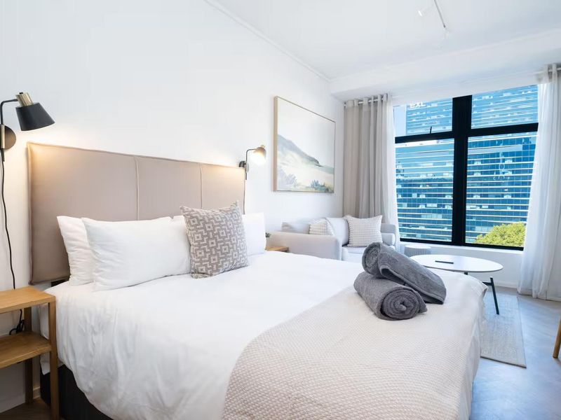 A well-lit bedroom featuring large windows with urban views, modern wall-mounted lighting, and light-toned hardwood flooring.