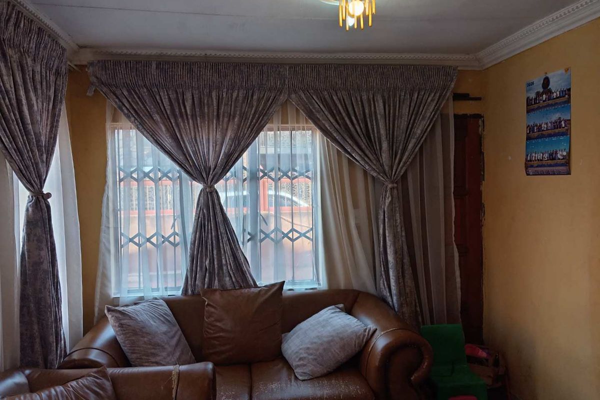 A living room featuring a window with security gate, decorative ceiling rose, and coving, requiring cosmetic updates to the wall finishes.