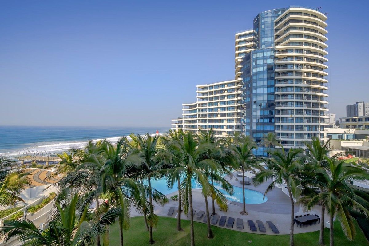 Aerial view of a modern high-rise apartment complex featuring a swimming pool, landscaped gardens, and direct proximity to the coastline.