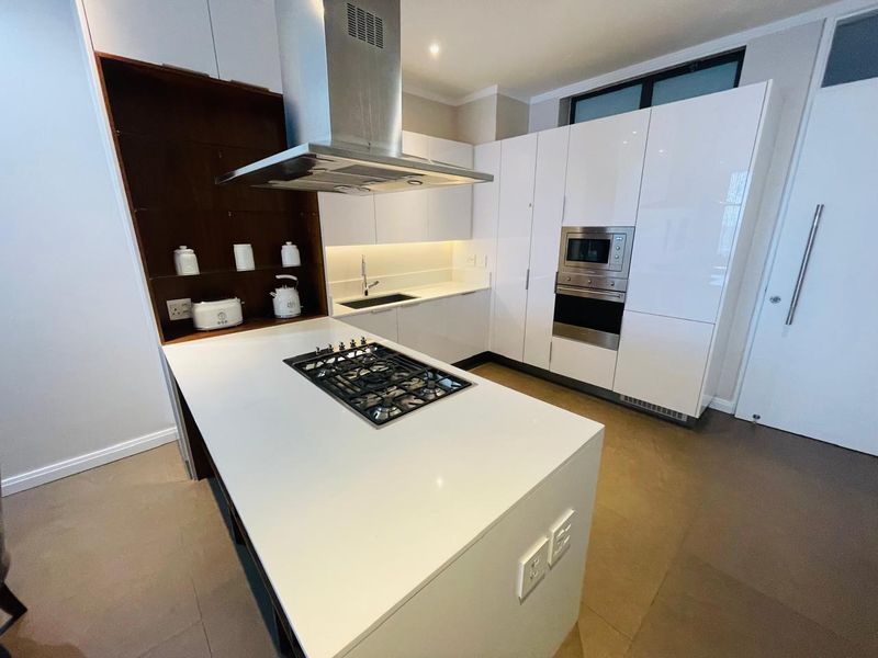 Modern kitchen featuring a central island with gas hob, high-gloss white cabinetry, integrated appliances, and sleek stone countertops with under-cabinet lighting.