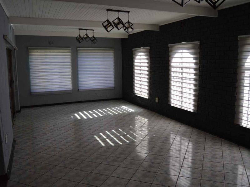 Spacious living area featuring tiled flooring, a painted brick feature wall, exposed beam ceiling, and ample natural light through multiple windows with fitted blinds.