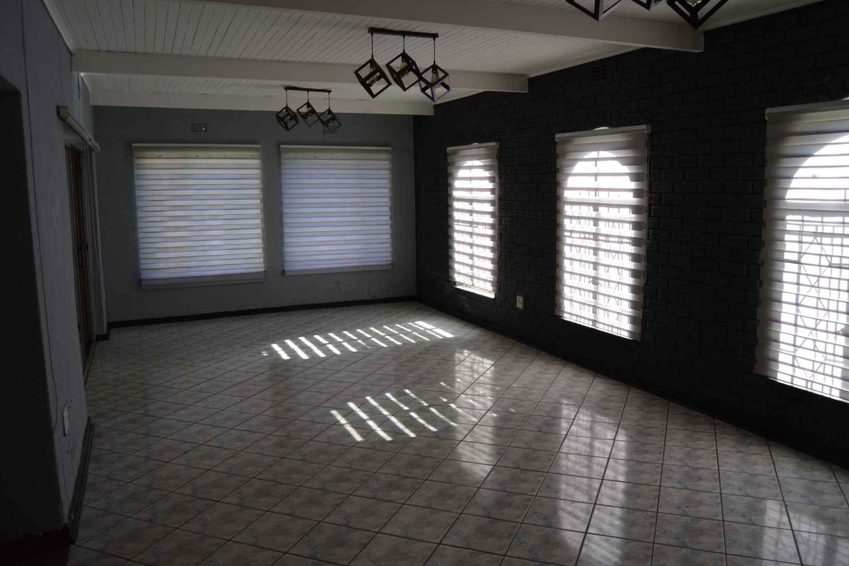 Spacious living area featuring tiled flooring, a painted brick feature wall, exposed beam ceiling, and ample natural light through multiple windows with fitted blinds.