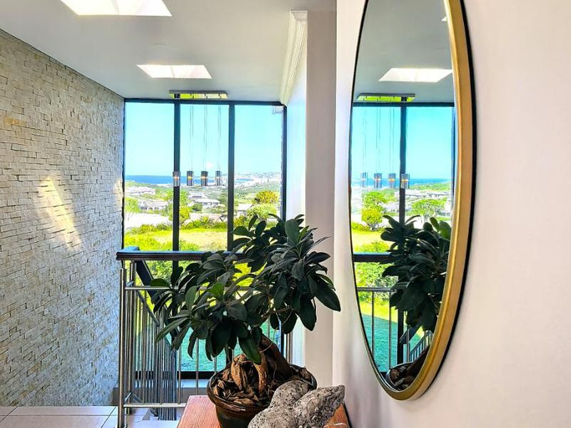 A bright hallway featuring a stone-clad feature wall, modern recessed lighting, and a metal balustrade overlooking a scenic outdoor view through large windows.