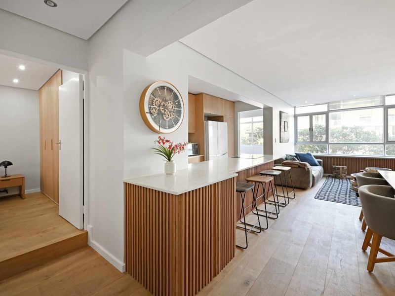 Modern open-plan living area featuring a stone-topped kitchen island with timber slat detailing, light wood flooring, and abundant natural light from large windows.