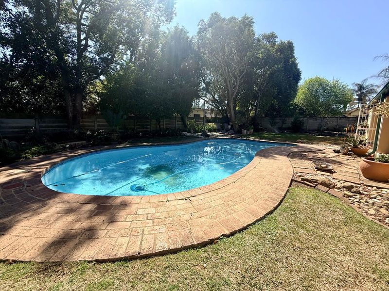 A backyard pool area featuring a curved swimming pool with brick coping, surrounded by a lawn and mature trees.