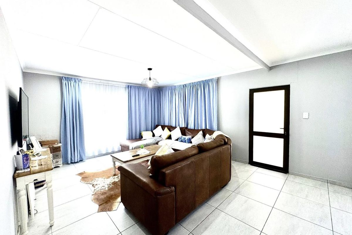 A bright living room featuring large format floor tiles, a central ceiling feature, and a glass-paneled door leading to an adjacent space.