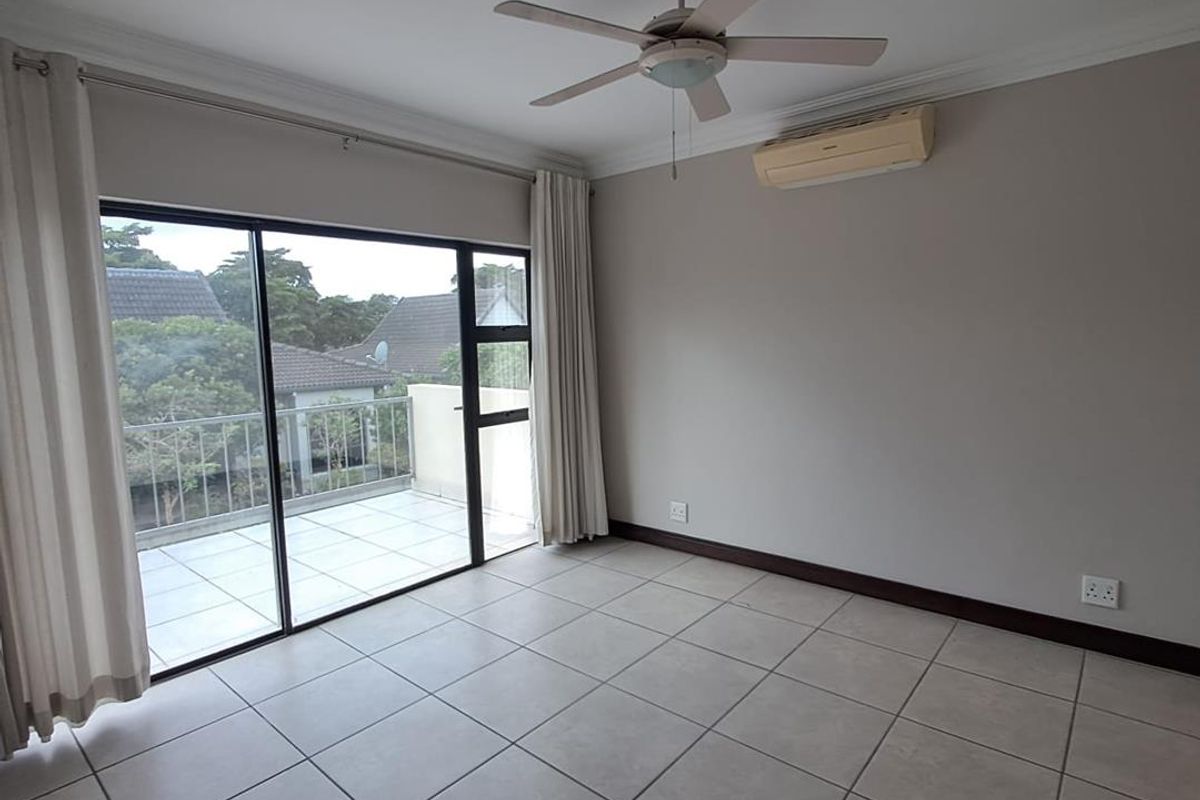 A bright, empty bedroom featuring tiled flooring, a ceiling fan, wall-mounted air conditioning, and large sliding glass doors opening onto a private balcony.