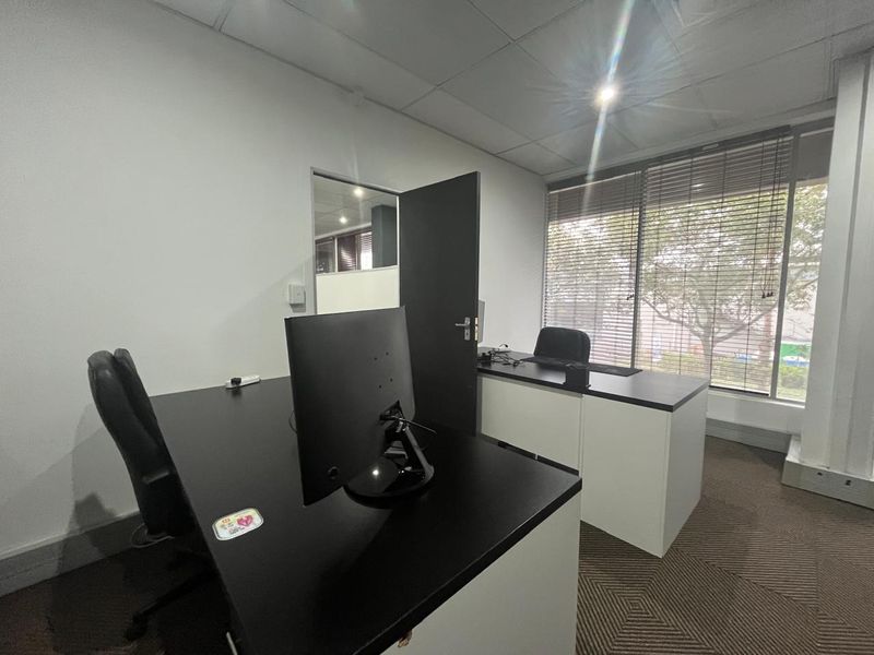 A functional office space featuring carpeted flooring, a suspended ceiling, and large windows with venetian blinds providing natural light.