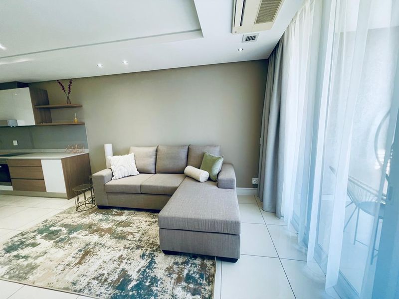 Modern open-plan living area featuring sleek kitchen cabinetry, built-in oven, recessed lighting, ducted air conditioning, and large sliding doors leading to a balcony.