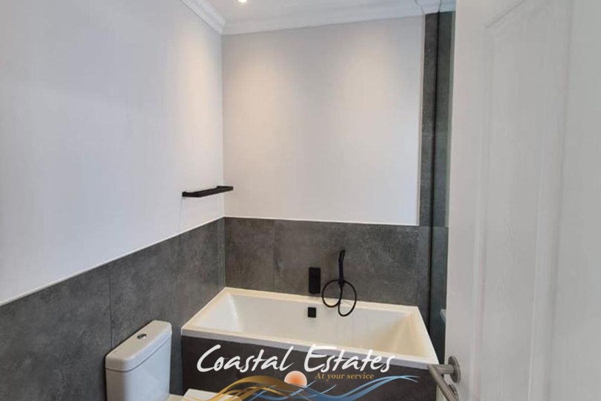 A modern bathroom featuring a built-in bathtub with matte black fixtures, a toilet, and contemporary grey wall tiling.