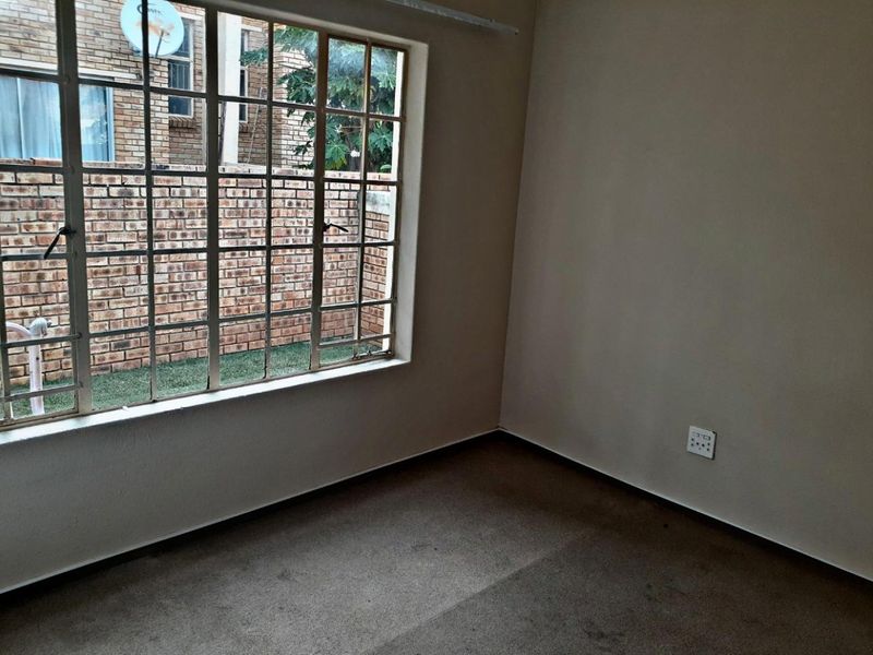 An empty bedroom featuring carpeted flooring and a large steel-framed window with security bars, offering a view of the exterior brick wall.