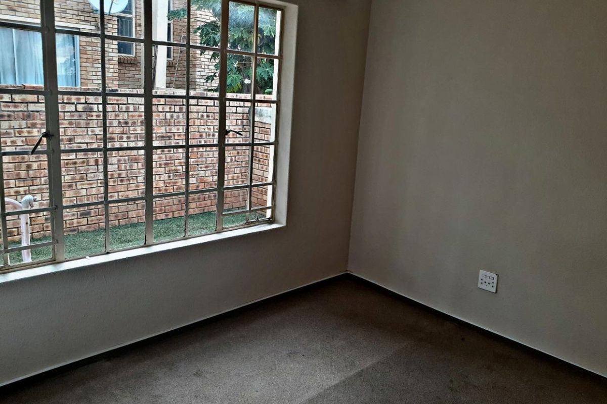 An empty bedroom featuring carpeted flooring and a large steel-framed window with security bars, offering a view of the exterior brick wall.