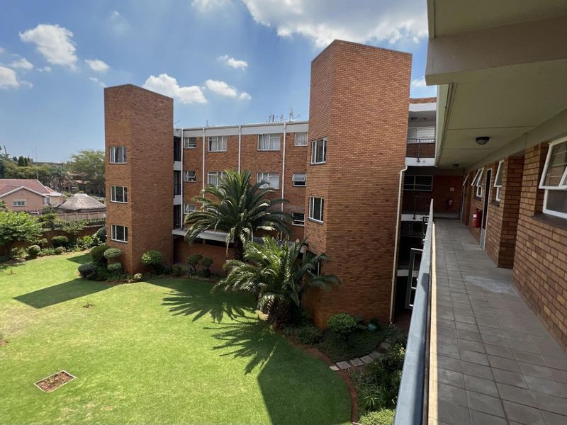 View of the apartment complex exterior featuring a brick facade, tiled access walkway, and a well-maintained communal garden area.
