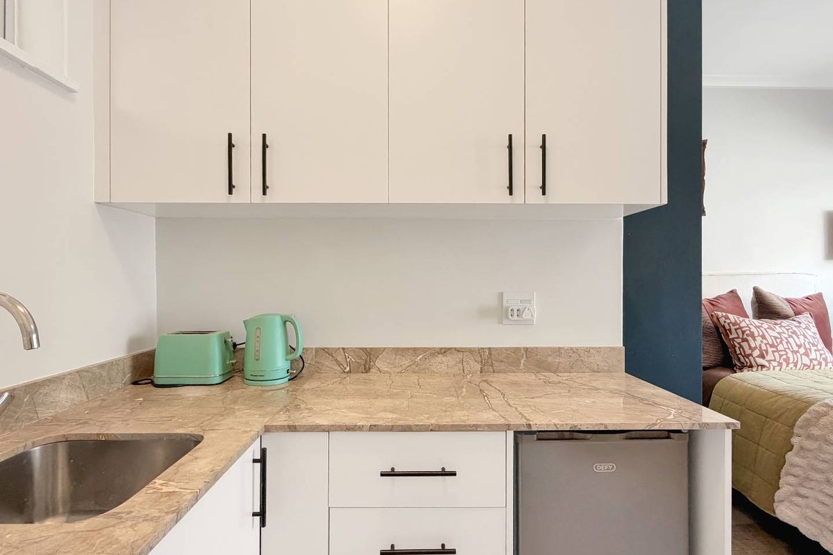 A compact, modern kitchen area featuring white cabinetry, stone countertops, and an integrated under-counter fridge, adjacent to a bedroom space.
