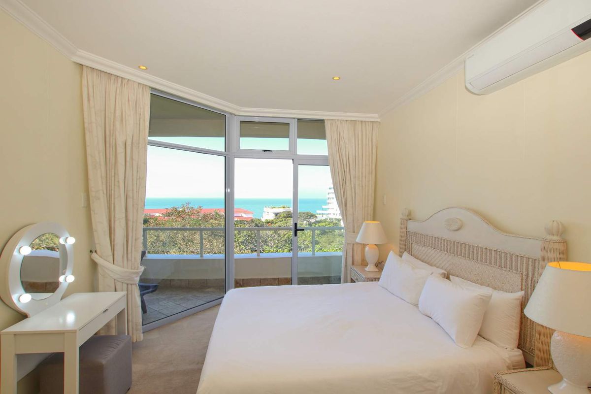 A bright bedroom featuring large glass sliding doors that open onto a private balcony with scenic ocean views and an integrated air conditioning unit.