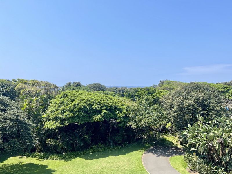 A scenic elevated view overlooking a lush green garden, mature trees, and a paved driveway with a distant horizon.