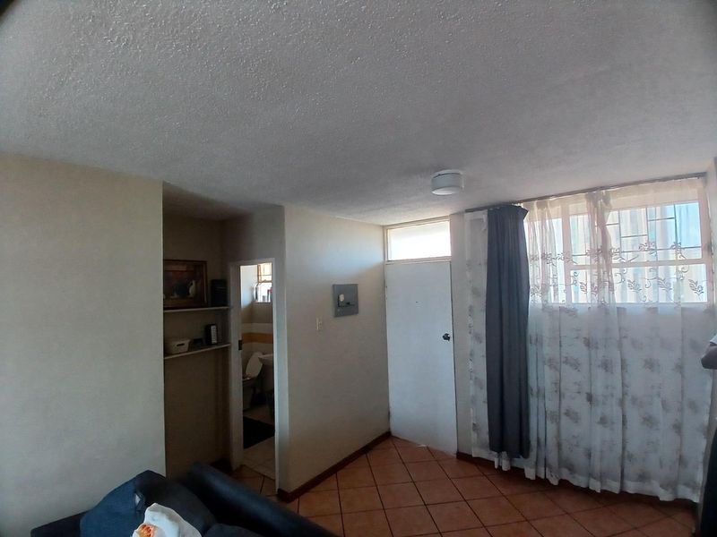 A living area featuring tiled flooring, built-in shelving, and access to an adjacent bathroom, with dated finishes and a security gate.