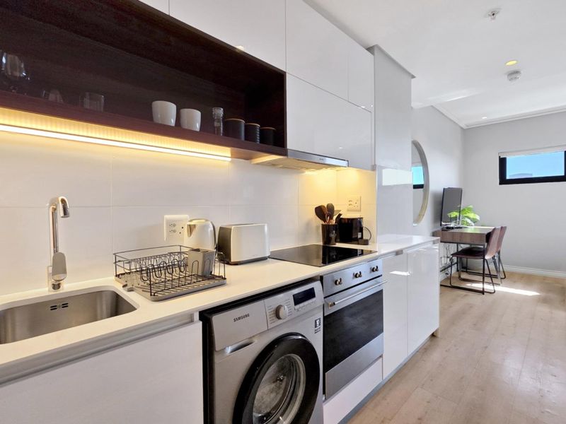 Modern open-plan kitchen featuring sleek white cabinetry, integrated appliances, under-cabinet lighting, and high-quality laminate flooring in a bright, contemporary apartment setting.