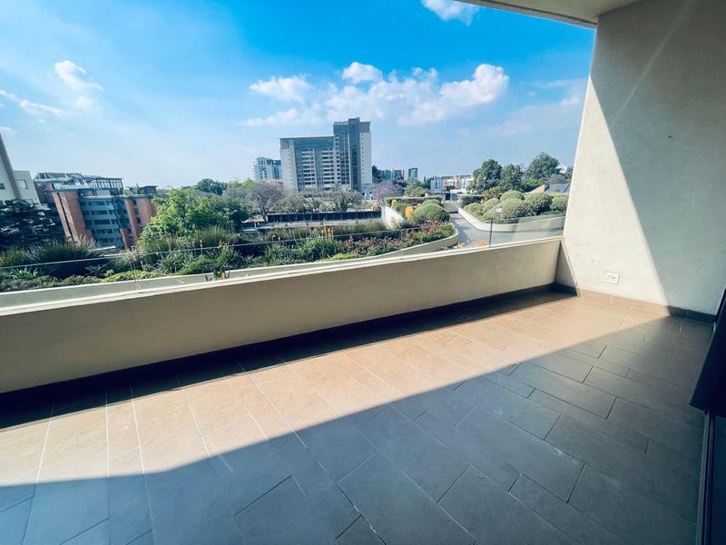 A spacious, modern balcony featuring tiled flooring and a sleek glass balustrade, offering elevated views of the surrounding urban landscape.