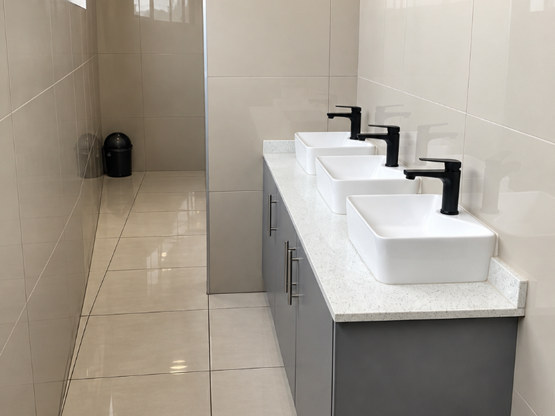 Modern commercial bathroom featuring a triple vanity with vessel sinks, matte black faucets, quartz countertop, and neutral floor-to-ceiling ceramic tiling.