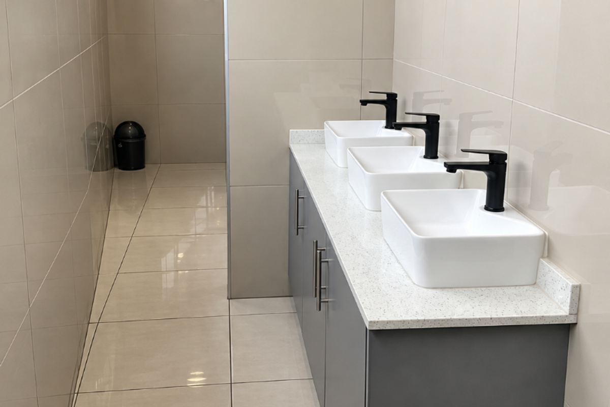 Modern commercial bathroom featuring a triple vanity with vessel sinks, matte black faucets, quartz countertop, and neutral floor-to-ceiling ceramic tiling.
