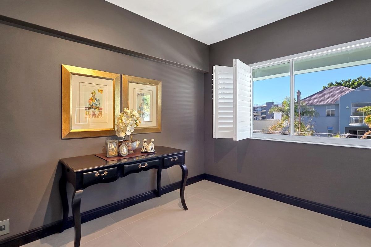 A well-maintained bedroom featuring neutral tiled flooring, dark accent walls, and white plantation shutters providing natural light and exterior views.