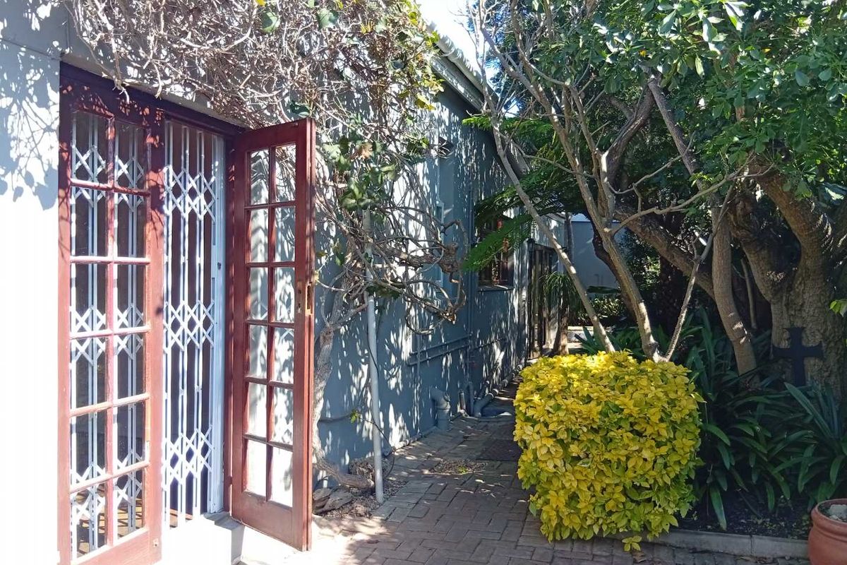 A rear exterior view featuring brick paving, mature garden landscaping, and french doors with integrated security gates providing access to the home.