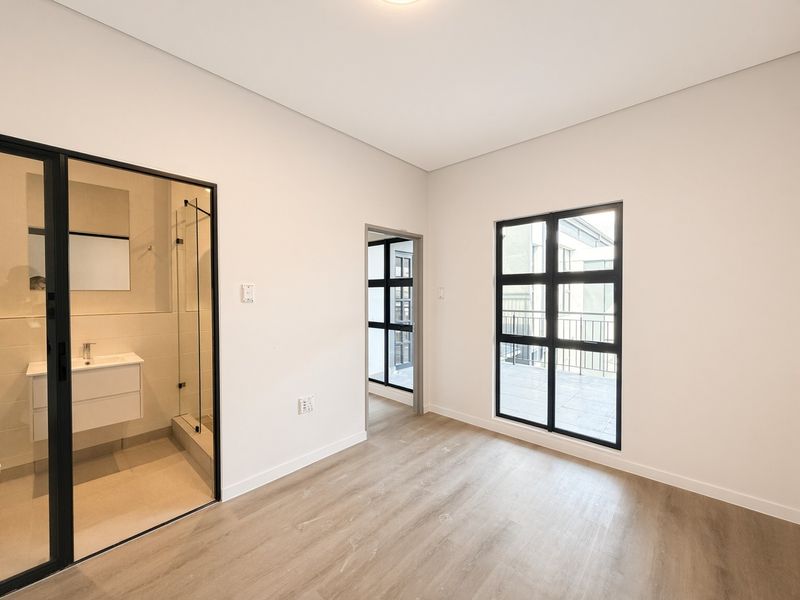 A bright, modern bedroom featuring laminate flooring, black-framed windows with balcony access, and a sleek en-suite bathroom with a glass-enclosed shower.