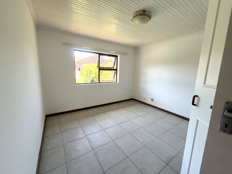 A bright, empty bedroom featuring neutral tiled flooring, a tongue and groove ceiling, and a window with security bars.