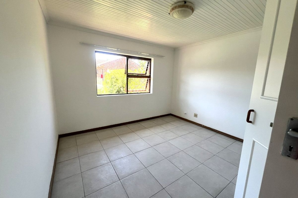 A bright, empty bedroom featuring neutral tiled flooring, a tongue and groove ceiling, and a window with security bars.
