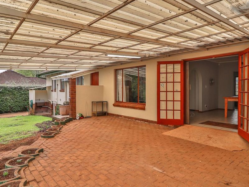 A covered outdoor patio area featuring brick paving, translucent roofing, and direct access to the interior through red-framed french doors.