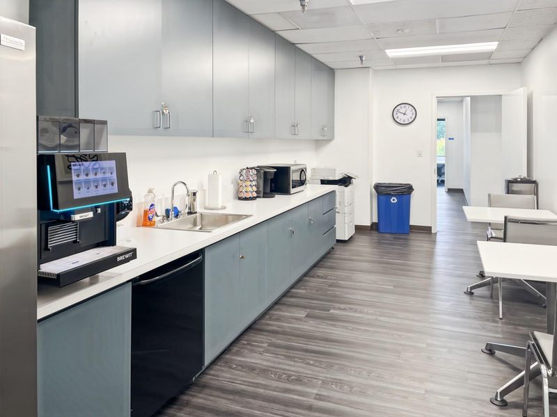 A clean and functional office kitchenette featuring grey cabinetry, a stainless steel sink, and durable laminate flooring.