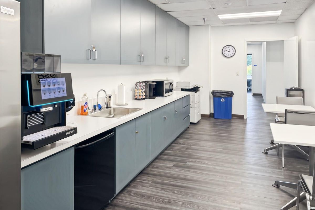 A clean and functional office kitchenette featuring grey cabinetry, a stainless steel sink, and durable laminate flooring.