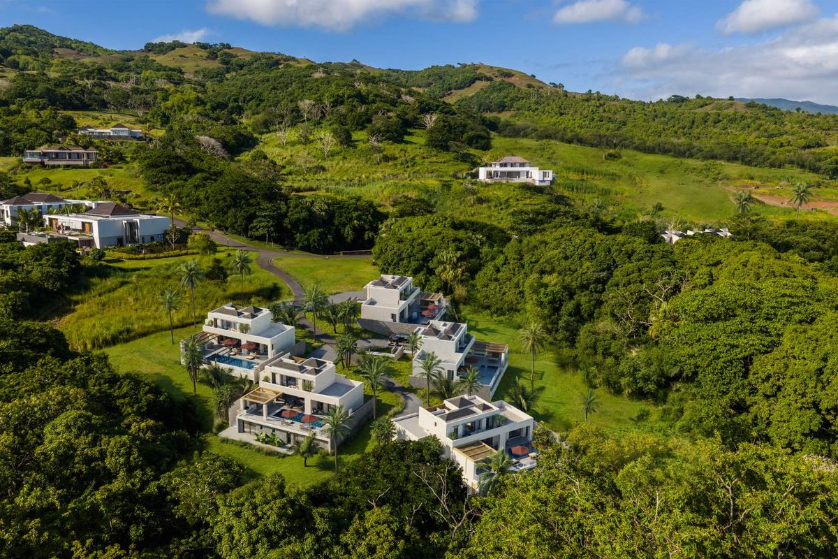 An aerial view of a modern residential estate featuring multiple contemporary villas nestled within a lush, green, hilly landscape.