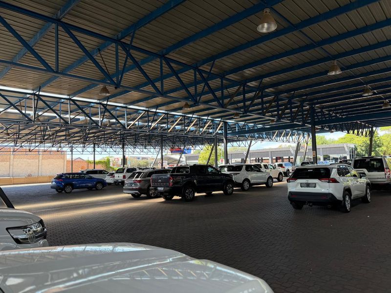 Large covered parking area with a robust steel truss roof structure and paved flooring, suitable for commercial vehicle display or customer parking.