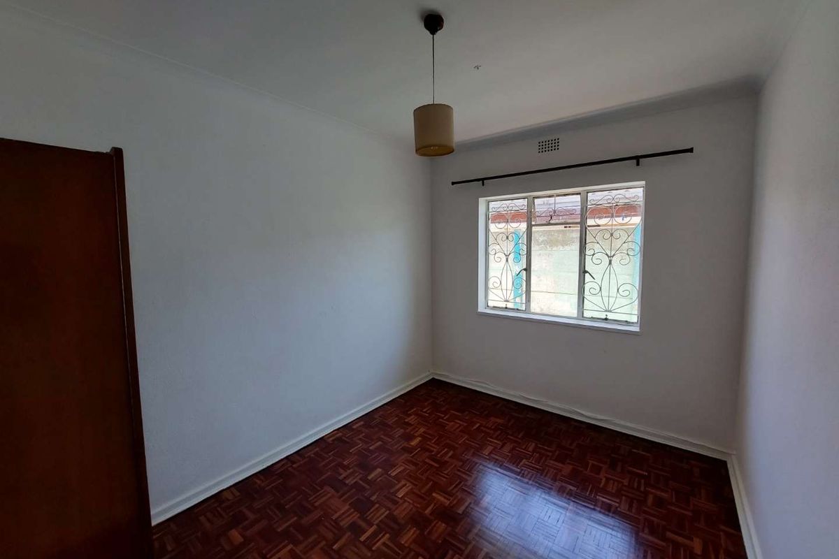 A bright, empty bedroom featuring classic parquet timber flooring, a single pendant light fixture, and a window fitted with decorative security bars.