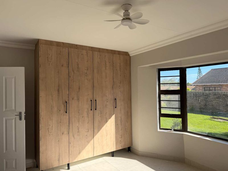 A bright, empty bedroom featuring modern wood-finish built-in cupboards, a ceiling fan, and tiled flooring with a view of the garden.