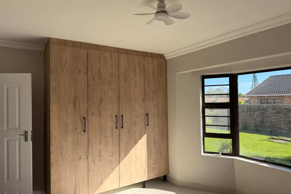 A bright, empty bedroom featuring modern wood-finish built-in cupboards, a ceiling fan, and tiled flooring with a view of the garden.