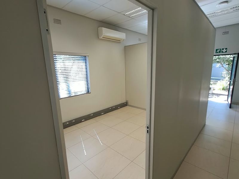 A clean, vacant office space featuring tiled flooring, suspended ceilings with recessed lighting, and a wall-mounted air conditioning unit.