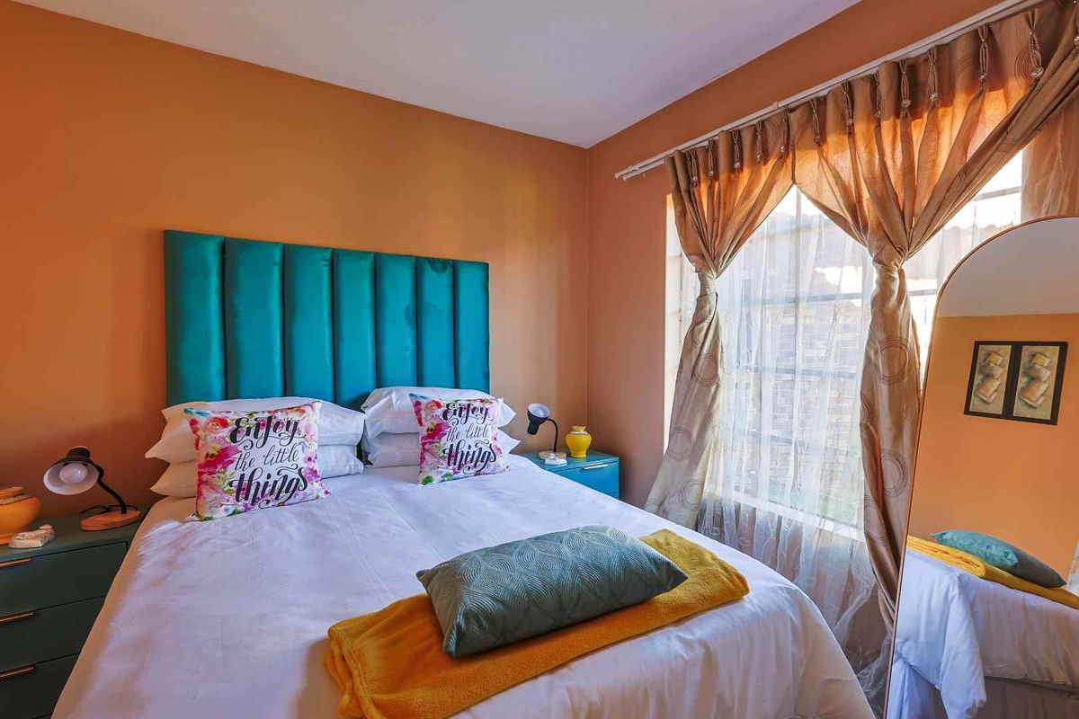 A cozy bedroom featuring a vibrant teal upholstered headboard, warm orange wall paint, and a large window with sheer curtains allowing natural light.