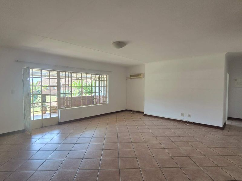 A spacious, empty living area featuring neutral tiled flooring, a wall-mounted air conditioning unit, and a large sliding glass door with security bars.