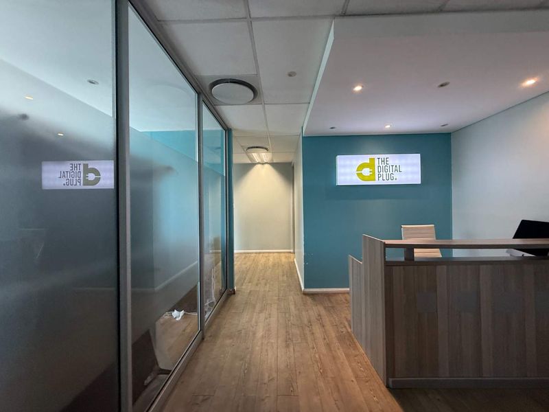 Modern office reception area featuring a glass partition wall, wood-look laminate flooring, recessed lighting, and a branded illuminated sign.