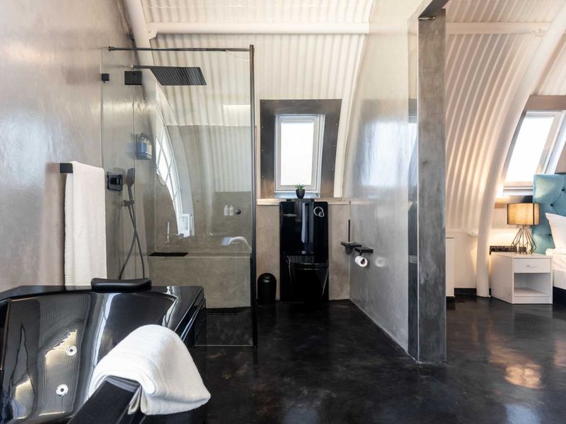 Modern open-plan bathroom featuring a sleek black bathtub, walk-in shower with rain head, black toilet, and polished concrete finishes under a unique corrugated ceiling.