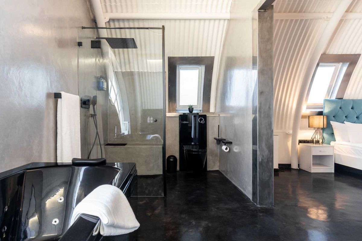 Modern open-plan bathroom featuring a sleek black bathtub, walk-in shower with rain head, black toilet, and polished concrete finishes under a unique corrugated ceiling.