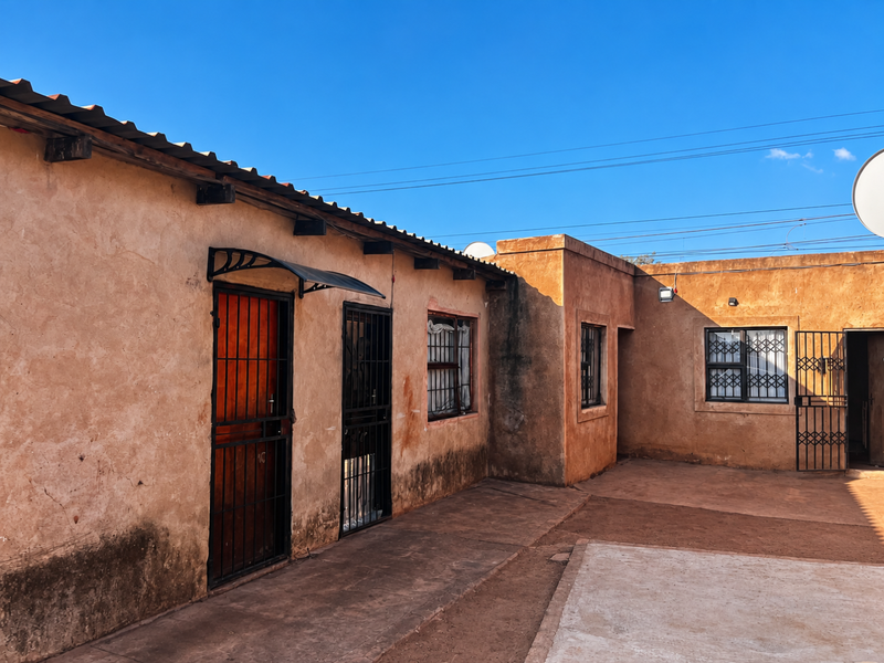 Rear exterior view of the property featuring a paved courtyard, security gates, and weathered wall finishes under a clear blue sky.