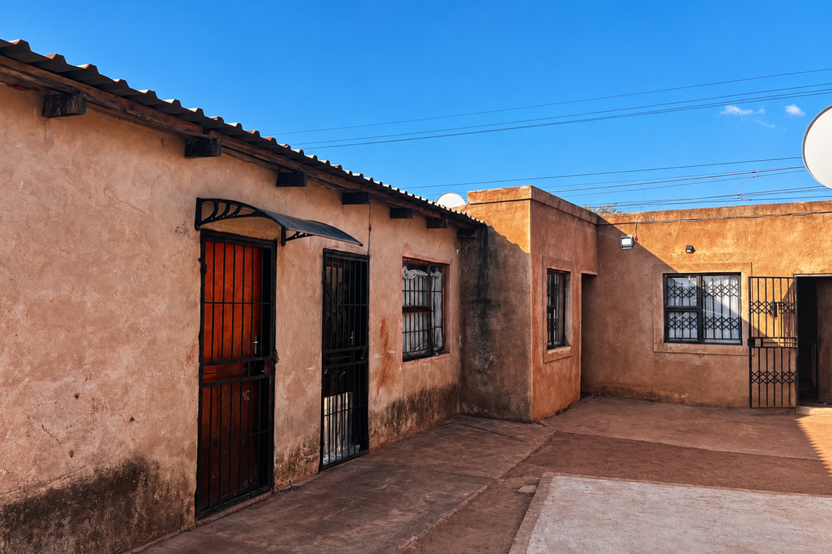 Rear exterior view of the property featuring a paved courtyard, security gates, and weathered wall finishes under a clear blue sky.
