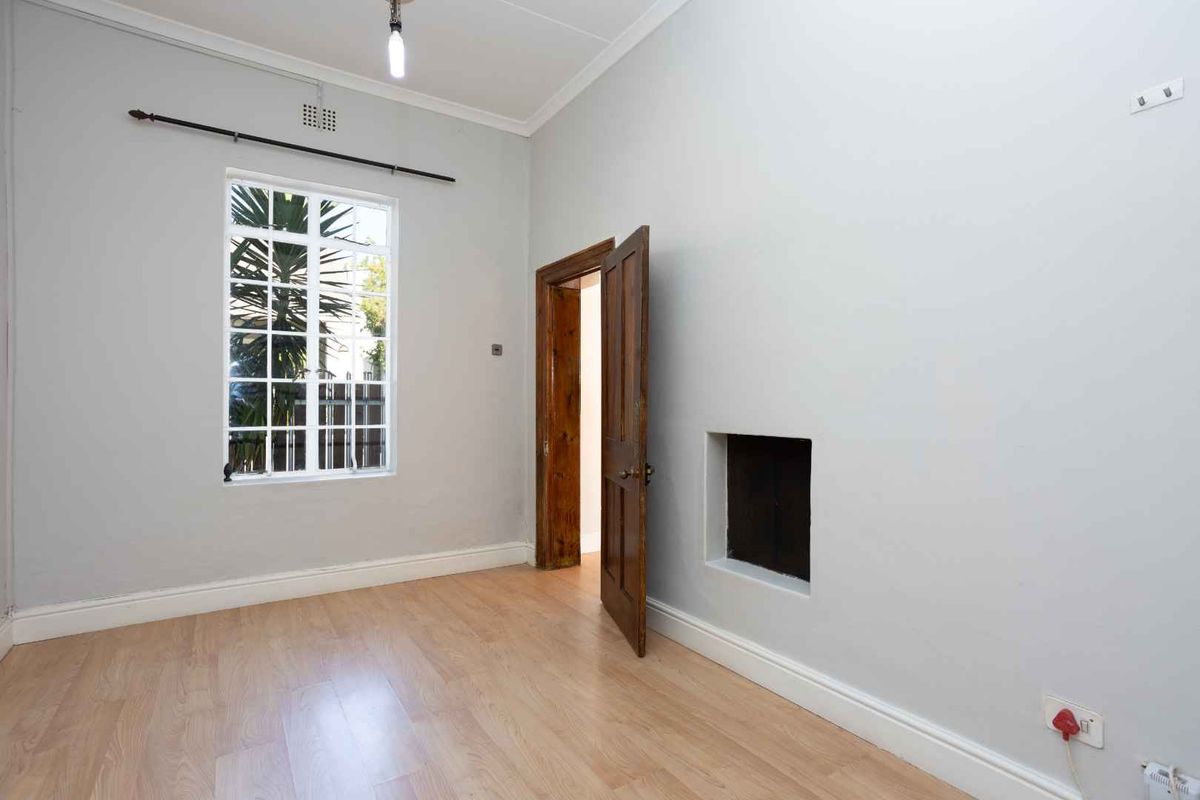 An empty bedroom featuring laminate flooring, a timber door, and a steel-framed window, currently showing a significant wall defect requiring repair.